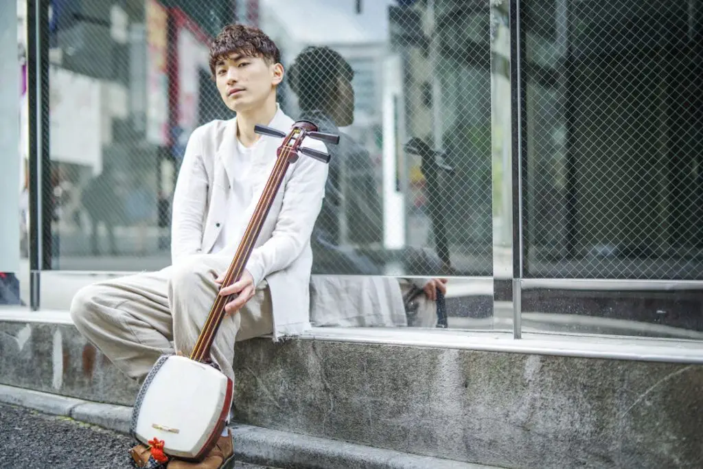 A Man in White Long Sleeves Sitting on the Street while Holding a Shamisen